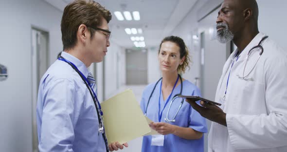 Diverse group of a female and two male doctors talking in hospital corridor holding tablet and files alt