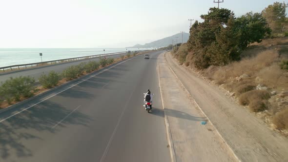 The Drone Follows a Motorcycle That Travels Along the Road Along the Sea Coast alt