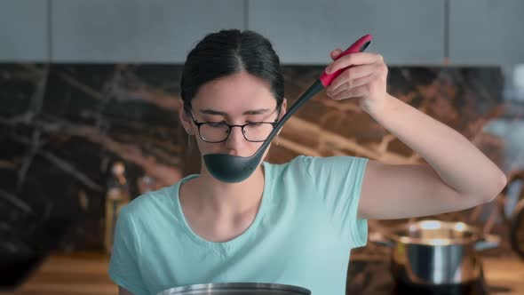 Happy Girl Tasting Soup on Spoon in Modern Kitchen and Smiling alt