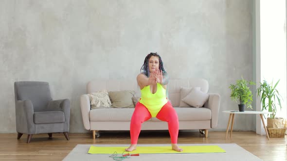 Fat Woman in Bright Sportswear with Braided Hairs Doing Squats Exercise at Home