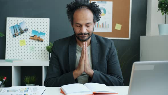 Pensive Middle Eastern Man Holding Hands in Namaste Gesture Sitting at Desk with Eyes Closed alt