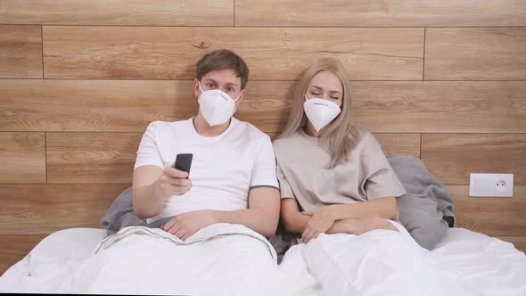 Portrait of Young Caucasian Couple in Medical Masks Sit on Bed Watching TV