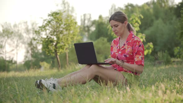 Woman Freelancer Working Remotely Sitting on Grass in Public Park Using Laptop