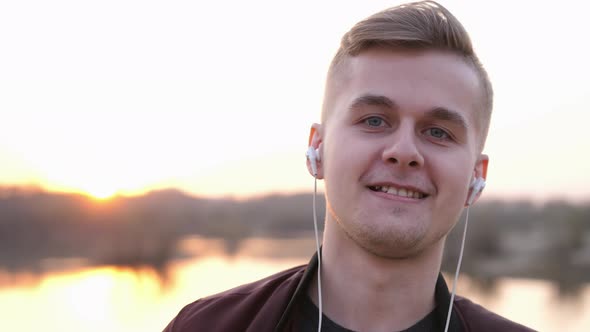 Young Smiling Handsome Man Listens to Music Looks at Camera on Beach During Sunset