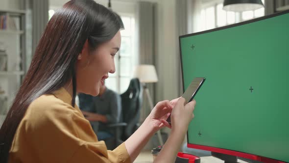 Asian Female Works And Using Mobile Phone With Mock Up Green Screen Computer Display In Office alt