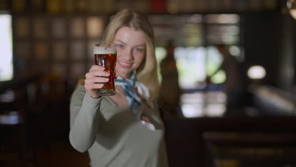 Happy Young Woman Toasting Stretching Beer Glass to Camera Smiling Standing in Pub Indoors alt