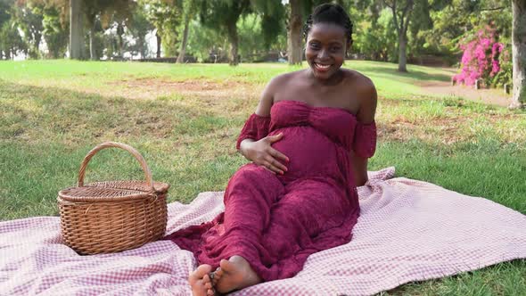 Young African woman caressing her pregnant belly while doing a picnic in park alt