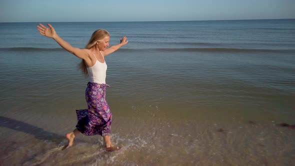 With arms outstretched, a slow motion clip of a mature woman splashing through the water on a beach alt