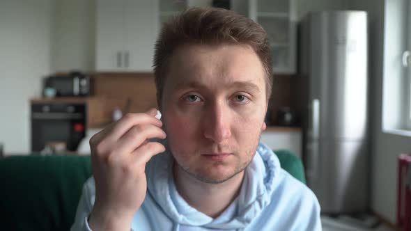 A Man Looks at the Camera at Home Inserts a Wireless Headset Into His Ears