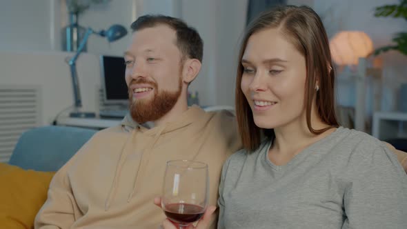Young Woman Watching TV with Boyfriend Drinking Red Wine and Feeding Man with Cheese Enjoying alt