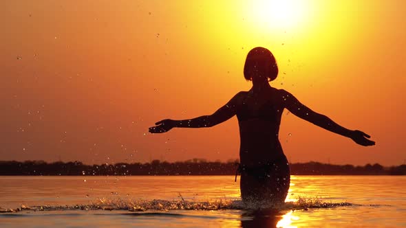 Silhouette of Woman at Sunset Raises Hands Up and Creating Splashes of Water. Slow Motion alt