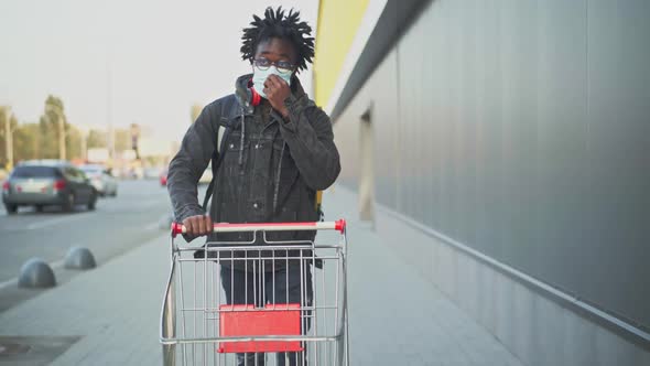 Portrait of Male Courier with Deadlocks in Covid Face Mask Pushing Shopping Cart To Shop alt