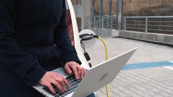 A Woman Sits on the Trunk of an Electric Car with a Laptop and Waits for the Car to Charge in the alt