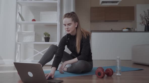 Girl Watching Training Videos with Sports Training at Home. Sitting on a Mat Near a Dumbbell alt
