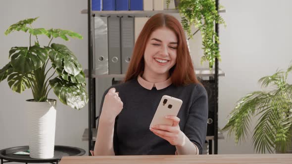 Portrait of happy surprised mixed race business woman celebrating victory on mobile phone at office.