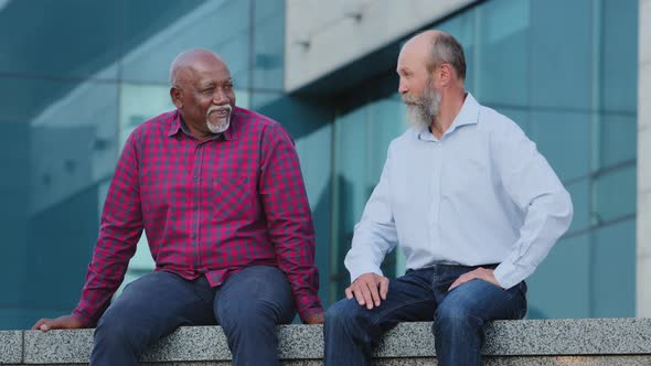 Two Diverse Bearded Elderly Male Friends in Casual Clothes Shirts and Jeans Sitting Outdoors alt