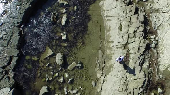 Aerial birds eye view shot of a young man running on a rocky ocean beach shoreline. alt