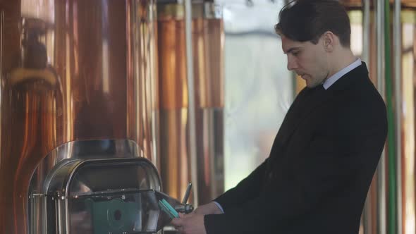Side View of Professional Caucasian Manager Standing at Brewing Equipment with Tablet Checking alt