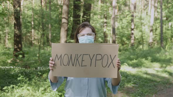 Woman in Mask Glasses Stands with Sign Monkeypox in Green Sunny Forest alt