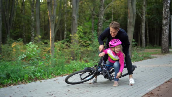 Young guy and girl teach children to ride a bike in the Park and fall. Two little girls in helmets