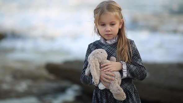 Little Sad Girl with Blond Hair Stands Against the Sea on a Very Windy Day