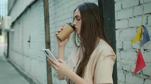 Pensive Young Woman Touching Smartphone Screen and Drinking to Go Coffee Outdoors in City Street alt