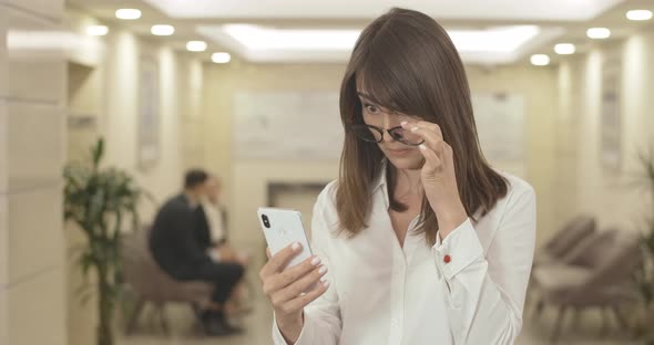 Portrait of Confused Young Brunette Woman in Eyeglasses Trying To Read on Smartphone Screen alt