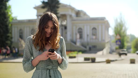 Portrait of Brunette Cute Woman Wearing Dress Smiling and Using Smartphone While Walking Through alt