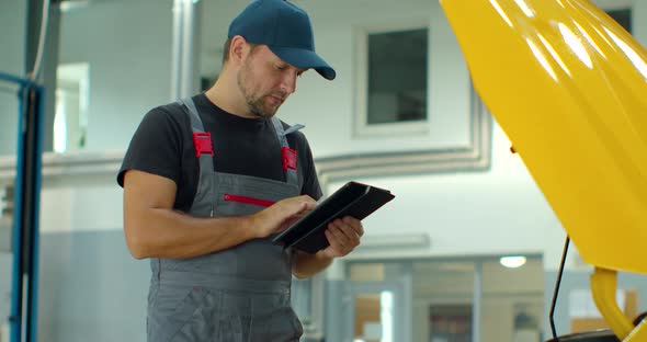 Mechanic Using a Digital Tablet While Doing Routine Maintenance Check-up on a Car in a Garage alt