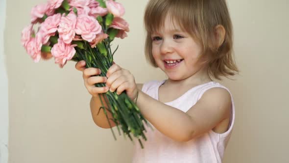 Little Girl Holds Bouquet of Flowers Pink Carnations Looks at Camera Smiles and Smells Flowers alt