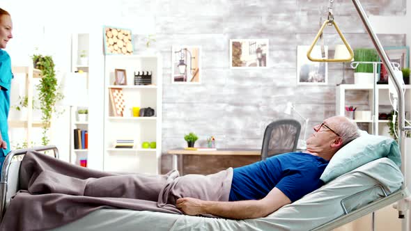 Female Nurse Helping an Old Disabled Sick Man Who Lies in Hospital Bed alt