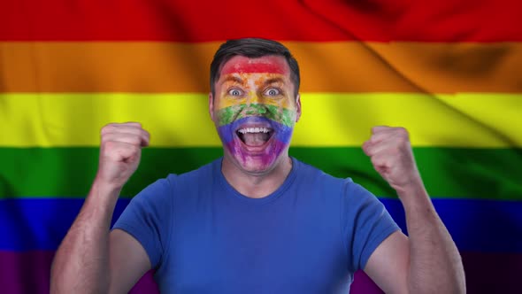 A screaming man with a face painted in the color of the LGBT flag.