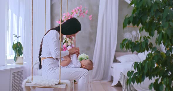 Young Beautiful Mother Sits on a Swing with Her Baby and Plays Talks and Smiles at Her Baby