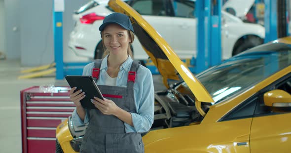 Female Car Mechanic Using Tablet Device in Big Garage with Cars. Beautiful Woman Using Tablet, Then alt