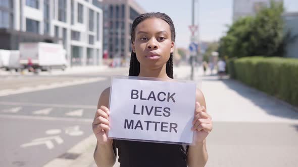 A Young Black Woman Holds a Black Lives Matter Sign in Front of the Camera - a Townscape alt