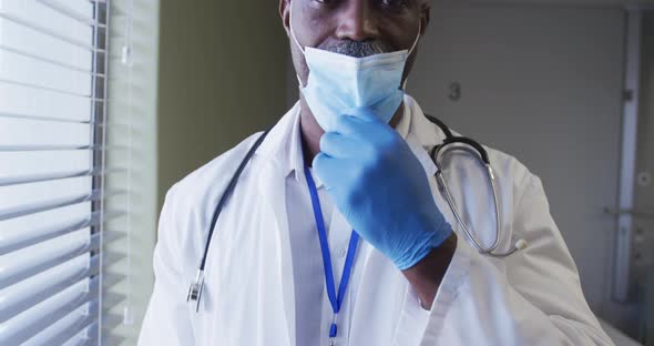Portrait of smiling african american male doctor wearing face mask standing in hospital room alt