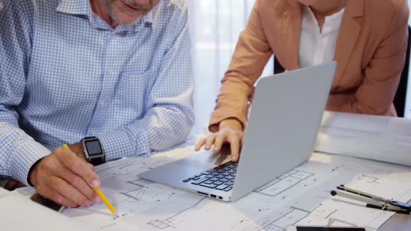 Businesswoman using laptop while coworker working on blueprint alt