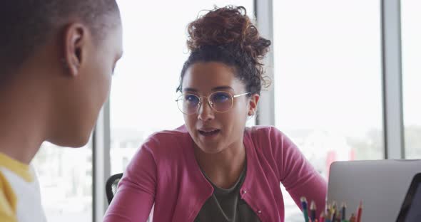 Two happy diverse creative businesswomen using laptops talking at desk in modern interior alt