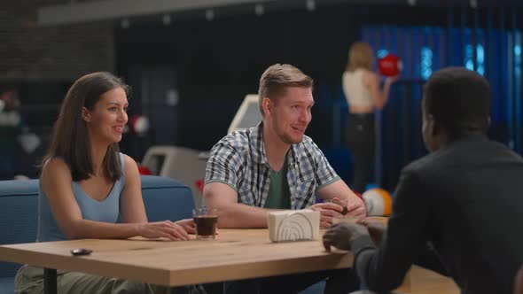 A Multiethnic Group of Friends Sits at a Table Together in a Bowling Alley and Talks alt