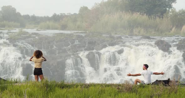 Woman is filming the gorgeous Victoria Falls and a man is sitting on a rock, 4k alt
