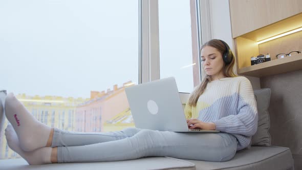 Young woman sitting by the window at home in headphones, working on a laptop. It's snowing outside. alt