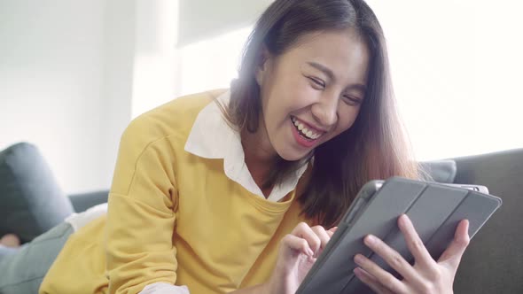 Asian woman using tablet watching video while wear sweater lying on sofa in living room. alt