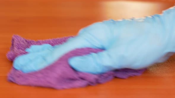 With a close palm, a hand in a medical glove wipes the surface of a wooden table with a rag alt