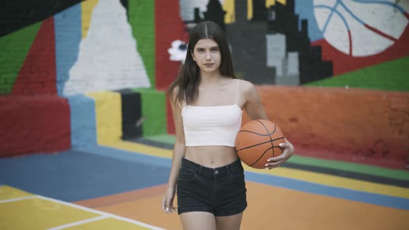 Portrait of Confident Brunette Young Sportswoman Walking with Basketball Ball Looking at Camera alt