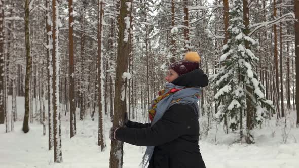 Smiling Young Woman Wearing Warm Clothes Shaking Tree and Getting Covered in Snow in Winter Forest alt
