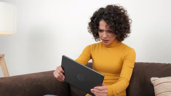 Curly Woman Emotionally Playing Video Game on Tablet Sitting on Couch at Home alt