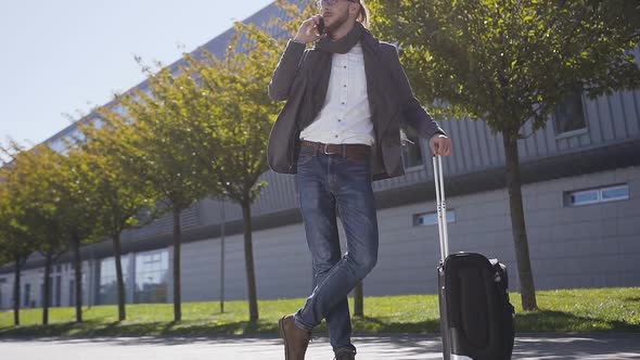 Young stylish man talking on his phone standing with suitcase  alt