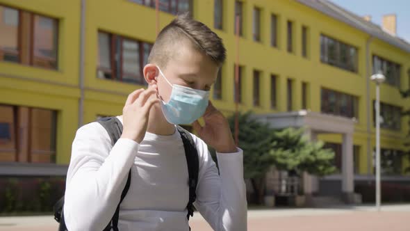 A Caucasian Teenage Boy Takes Off a Face Mask and Looks at the Camera  a School in the Background