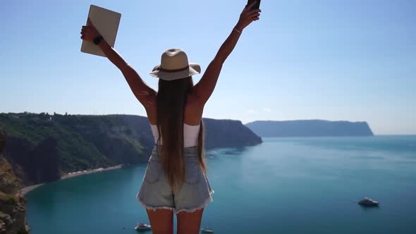 Digital Nomad Woman in the Hat a Business Woman with a Laptop Stands on the Rocks By the Sea During alt