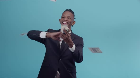 Black Man in a Strict Suit Smiling Throws a Pack of Dollar Bills in the Studio on Blue Background alt